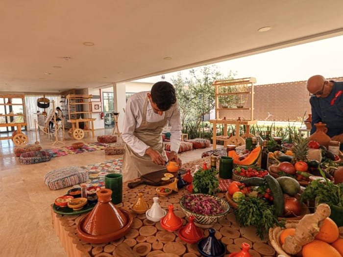 A cook preparing tagines surrounded by baskets of Moroccan produce and spices.